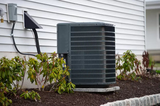 Outdoor AC condenser unit installed beside a house with shrubs and landscaping